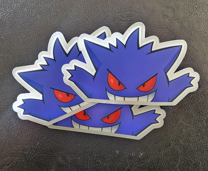 May include: Three layered stickers featuring a purple and blue cartoon ghost with red eyes and a wide grin. The ghost is shown with its arms outstretched.