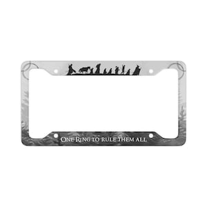 Puede incluir: Marco de matrícula con un diseño en blanco y negro que presenta una silueta de figuras y el texto "ONE RING TO RULE THEM ALL". El marco tiene un fondo gris con una escena de bosque.