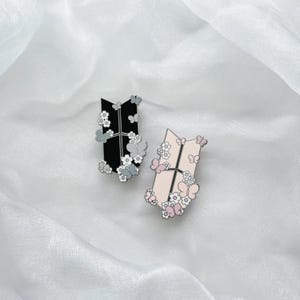 Puede incluir: Dos pines de esmalte con un logotipo estilizado con flores y mariposas. Un pin es negro con flores y mariposas blancas, y el otro es rosa con flores y mariposas blancas.