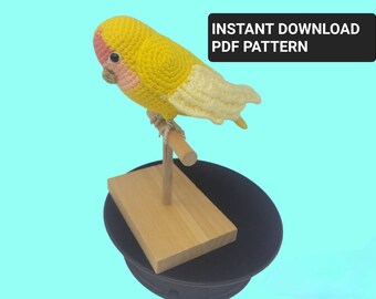 May include: A crocheted yellow and pink bird perched on a wooden stand. The bird has a white wing and is sitting on a black surface. The text "INSTANT DOWNLOAD PDF PATTERN" is in a black rectangle at the top of the image.