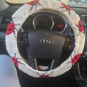 May include: A crocheted steering wheel cover in an off-white color featuring a repeating pattern of red ladybugs with black spots. The cover is fitted over a black Kia steering wheel with integrated control buttons and a central Kia logo.