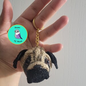 Könnte beinhalten: Ein gehäkelter Mops-Schlüsselanhänger mit schwarzen Ohren und Nase. Der Schlüsselanhänger ist an einer goldenen Kette und einem Ring befestigt. Ein runder Aufkleber mit einem blauen Vogel und dem Text "Dreams of Thread" befindet sich im Hintergrund.