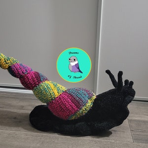 May include: A colorful crocheted snail with a black fuzzy shell. The snail is curled up with its head and tentacles sticking out. The snail is on a wooden floor. The image includes a logo for "Dreams of Thread".