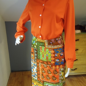 May include: A bright orange long-sleeved shirt with a collared neckline and button closure. The shirt is paired with a colorful patchwork skirt featuring paisley and floral patterns in shades of brown, green, blue, and yellow.