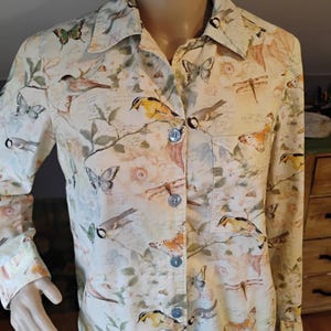 May include: A button-down shirt with a cream-colored background, adorned with a pattern of birds, butterflies, and dragonflies. The long-sleeved shirt features a collared neckline and light blue buttons. The design includes floral and botanical elements.