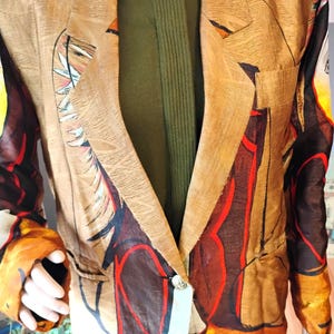 May include: Tan blazer with an abstract design in shades of brown, red, and orange. The blazer has a notched lapel and a single button closure. A green sweater is visible underneath.