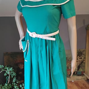 May include: A teal dress with a white collar and trim, short sleeves, and a white belt. The dress has a pleated skirt and a pocket with white trim. The dress is displayed on a mannequin.