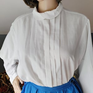 May include: A white long-sleeved blouse featuring a rounded collar with delicate embroidery and vertical pleats running down the front chest. The blouse is paired with a bright blue skirt that has a pleated waistband and a side button closure.
