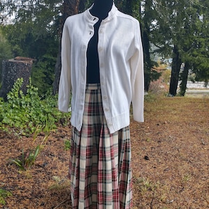 May include: A white cardigan sweater with a button closure and long sleeves, paired with a plaid skirt with red, green, and white colors.