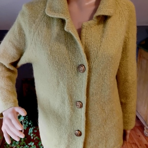 May include: A light green knitted jacket with a collared neckline and three decorative buttons. The jacket has long sleeves and a textured appearance. The buttons are a bronze color. The jacket is displayed on a mannequin.