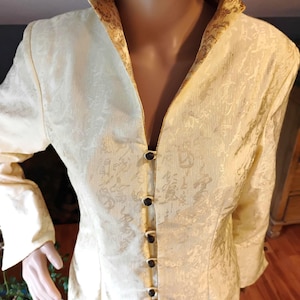 May include: Cream-colored jacket with a subtle floral pattern. The jacket features a mandarin collar and is fastened with dark buttons. The collar and lapel have a gold-colored decorative trim. The sleeves are long and the fabric has a slight sheen.