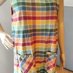 May include: A sleeveless apron with a colourful plaid pattern in shades of red, orange, yellow, green, and blue. The apron has two large pockets and a white lace trim at the bottom.