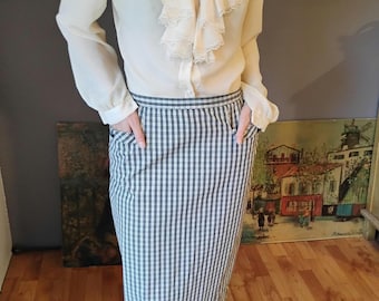 May include: Cream-colored blouse with a ruffled collar and cuffs, paired with a black and white checkered pencil skirt. The skirt has pockets and falls to the knees. The outfit is classic and stylish.
