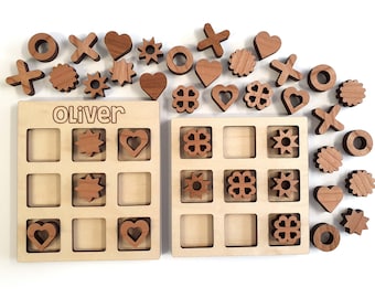 May include: A wooden tic-tac-toe game with two boards and extra pieces. The boards are made of light wood and have a grid pattern. The pieces are made of dark wood and have different shapes, including hearts, stars, and circles. The name "Oliver" is engraved on one of the boards.