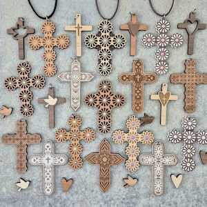 May include: A collection of wooden cross pendants in various designs, including floral, heart, and geometric patterns. Some crosses are adorned with small embellishments and hang on black or brown cords. Additional wooden cutouts of birds and hearts are scattered around.