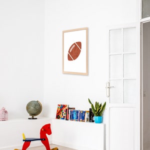 Puede incluir: Una impresi&oacute;n enmarcada de una pelota de f&uacute;tbol marr&oacute;n sobre un fondo blanco. La impresi&oacute;n est&aacute; colgada en una pared blanca en una habitaci&oacute;n con un caballo de balanc&iacute;n, un globo terr&aacute;queo y una estanter&iacute;a.