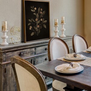 K&ouml;nnte beinhalten: Ein gerahmter botanischer Druck eines dunkelgr&uuml;nen, belaubten Zweigs mit zarten rosa Bl&uuml;ten auf schwarzem Hintergrund. Der Druck h&auml;ngt an einer Wand &uuml;ber einem Holzbuffet mit einem goldenen Metallbuchstaben S und zwei wei&szlig;en Kerzenhaltern.