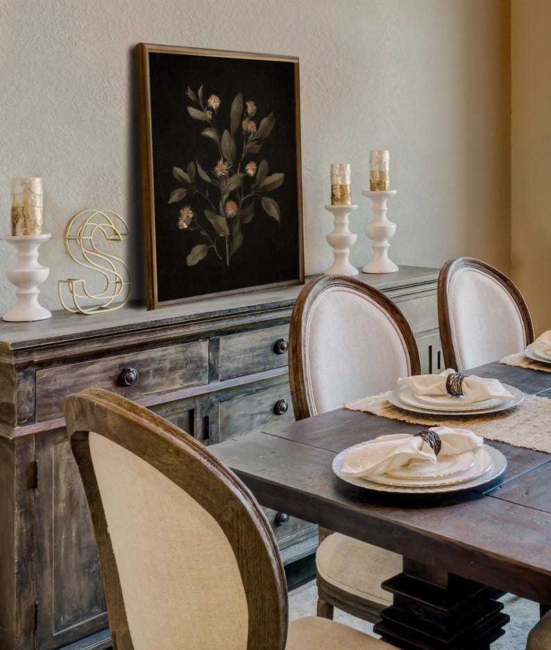 K&ouml;nnte beinhalten: Ein gerahmter botanischer Druck eines dunkelgr&uuml;nen, belaubten Zweigs mit zarten rosa Bl&uuml;ten auf schwarzem Hintergrund. Der Druck h&auml;ngt an einer Wand &uuml;ber einem Holzbuffet mit einem goldenen Metallbuchstaben S und zwei wei&szlig;en Kerzenhaltern.