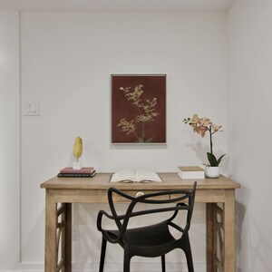 Op de afbeelding: Een houten bureau met een zwarte stoel, een ingelijst botanisch drukwerk en een orchidee in een pot. Het bureau heeft een lade en een boek erop.