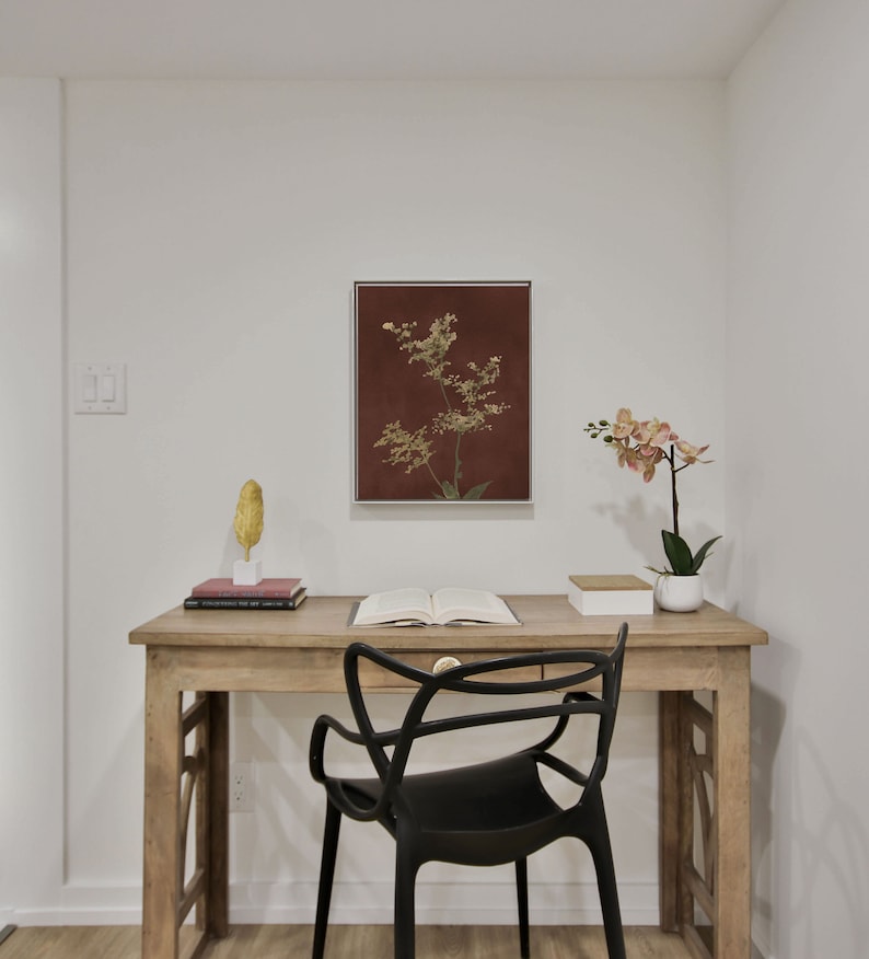 Op de afbeelding: Een houten bureau met een zwarte stoel, een ingelijst botanisch drukwerk en een orchidee in een pot. Het bureau heeft een lade en een boek erop.