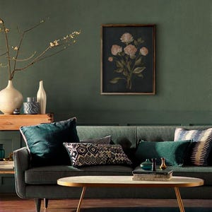 May include: A living room scene with a dark green sofa, throw pillows, and a wooden framed floral print. A gold-toned coffee table sits in front of the sofa. Decorative vases and a wooden box are on a side table. The wall is painted a dark green.