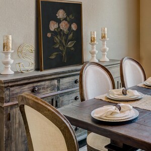 May include: A dining room scene featuring a dark wooden table set for a meal. The table is surrounded by chairs with cream-colored upholstered backs. A framed floral print hangs above a wooden sideboard, with white candle holders and a gold wire letter 'S'.