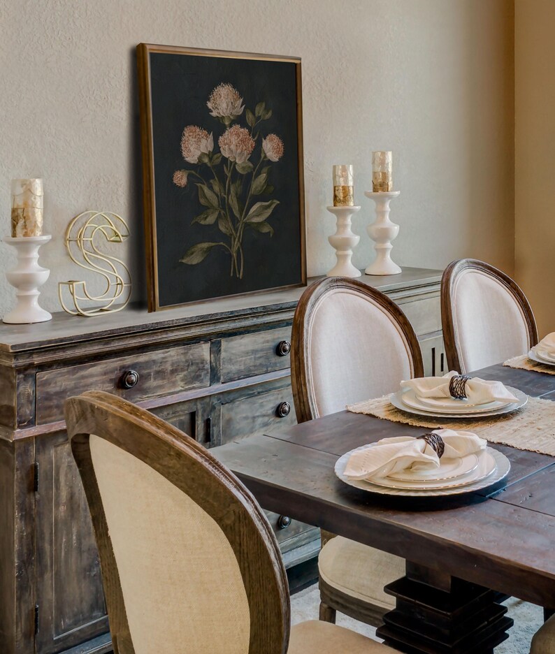 May include: A dining room scene featuring a dark wooden table set for a meal. The table is surrounded by chairs with cream-colored upholstered backs. A framed floral print hangs above a wooden sideboard, with white candle holders and a gold wire letter 'S'.