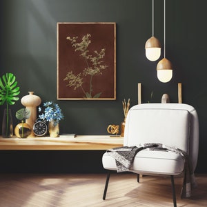 Op de afbeelding: Een witte fauteuil met een zwart metalen frame en een grijs-witte deken. De stoel staat voor een donkergroene muur met een ingelijst afdruk van een bruine en groene botanische illustratie. Twee houten hanglampen hangen aan het plafond.