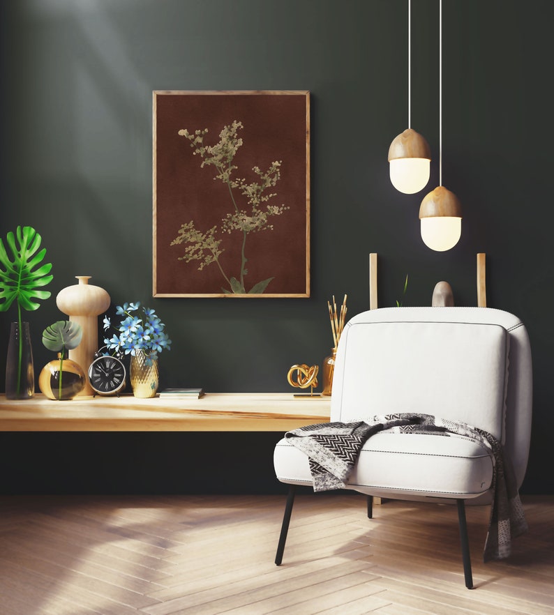 Op de afbeelding: Een witte fauteuil met een zwart metalen frame en een grijs-witte deken. De stoel staat voor een donkergroene muur met een ingelijst afdruk van een bruine en groene botanische illustratie. Twee houten hanglampen hangen aan het plafond.