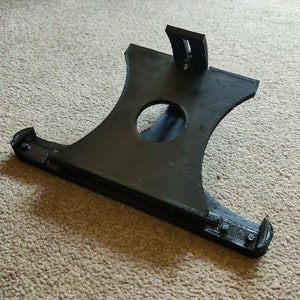 May include: A black 3D printed phone stand with a circular cutout in the center. The stand has a raised section on the back for propping up the phone.