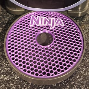 Op de afbeelding: Een paarse, honingraatvormige basis met het woord "NINJA" in witte letters. De basis is ontworpen voor een keukenapparaat.