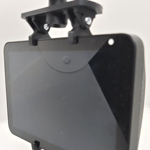 May include: A black tablet secured by a black mounting bracket. The tablet is held in place by two small clips at the bottom and a cover at the top. The bracket is attached to a larger black mounting system.