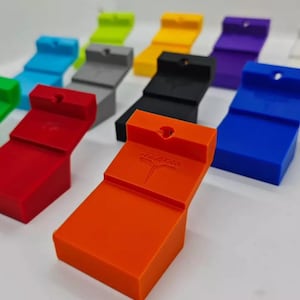 May include: A collection of 10 colorful 3D printed wall mounts, each with a unique color and a small hole at the top. The mounts are arranged in a staggered pattern, with the orange mount in the foreground. The orange mount has the word "remarkable" printed on it in white.
