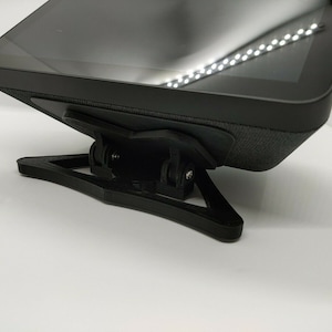 May include: A black stand with a triangular base supports a black tablet device. The stand is made of plastic and has a textured surface. The tablet is angled slightly upward.