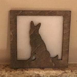 May include: A square, wooden frame with a cutout silhouette of a dog, likely a German Shepherd, sitting upright. The dog is brown and the background is white. The frame has a textured, stone-like appearance.