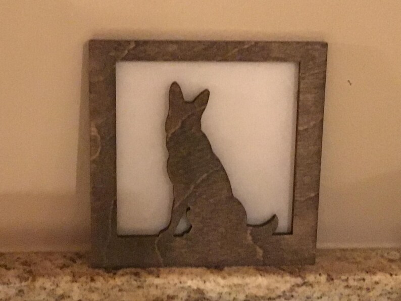 May include: A square, wooden frame with a cutout silhouette of a dog, likely a German Shepherd, sitting upright. The dog is brown and the background is white. The frame has a textured, stone-like appearance.
