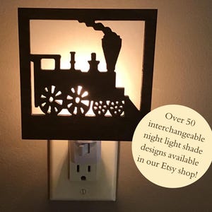 May include: A black night light with a train silhouette design. The train has detailed wheels and a smokestack. The night light is plugged into a wall outlet. Text on the image says "Over 50 interchangeable night light shade designs available in our Etsy shop!"