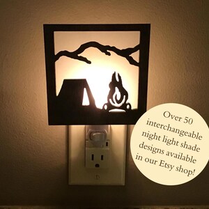 May include: A black metal night light with a white silhouette of a tent and campfire in front of a mountain range. The night light is plugged into an electrical outlet. The text "Over 50 interchangeable night light shade designs available in our Etsy shop!" is written in a circle below the night light.