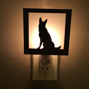 May include: A black silhouette of a dog, likely a German Shepherd, sits inside a square frame. The frame is mounted on a night light that plugs into a wall outlet. The background glows with a warm, soft light.