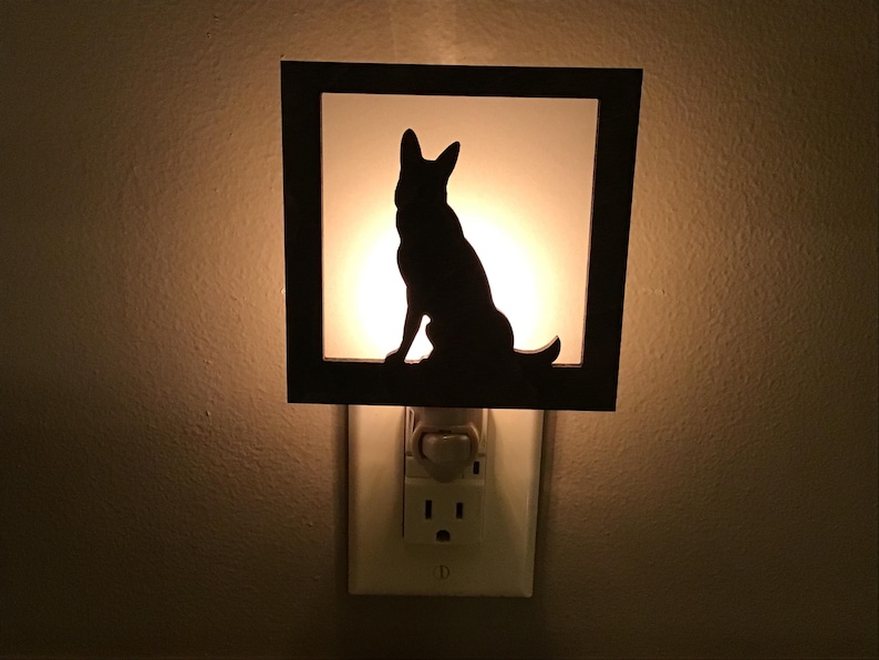 May include: A black silhouette of a dog, likely a German Shepherd, sits inside a square frame. The frame is mounted on a night light that plugs into a wall outlet. The background glows with a warm, soft light.