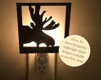 May include: A wooden moose silhouette night light plugged into an electrical outlet. The light is on and illuminates the moose shape. Text on the image reads "Over 50 interchangeable night light shade designs available in our Etsy shop!"