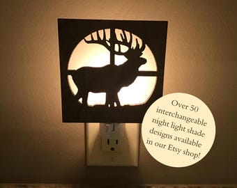 May include: A decorative night light with a silhouette of an elk inside a circular cutout. The elk is set against a bright background, and the night light is plugged into a wall outlet. The night light has a dark brown frame. Text on the image says "Over 50 interchangeable night light shade designs available in our Etsy shop!"