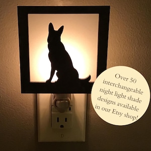 May include: Black silhouette of a dog inside a square night light. The night light is plugged into a wall outlet. The text on the image says "Over 50 interchangeable night light shade designs available in our Etsy shop!"