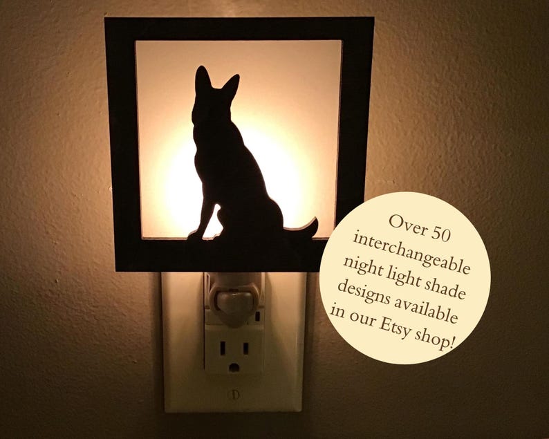 May include: Black silhouette of a dog inside a square night light. The night light is plugged into a wall outlet. The text on the image says "Over 50 interchangeable night light shade designs available in our Etsy shop!"