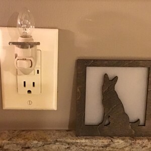 May include: A decorative wooden frame with a silhouette of a dog, next to a night light plugged into a wall outlet. The night light has a small, clear bulb. The frame is gray and the wall is beige.