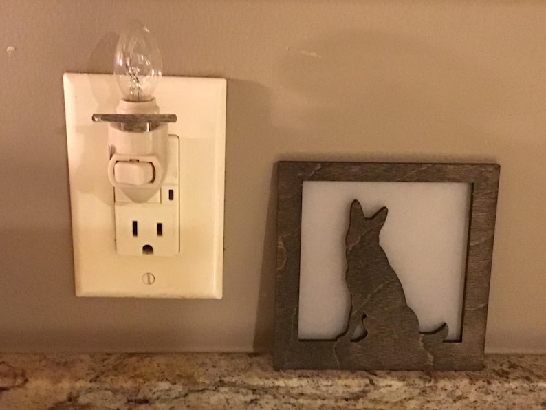 May include: A decorative wooden frame with a silhouette of a dog, next to a night light plugged into a wall outlet. The night light has a small, clear bulb. The frame is gray and the wall is beige.