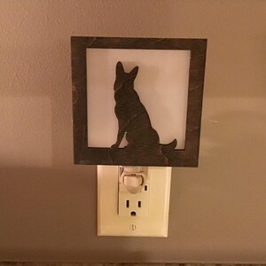 May include: A decorative night light featuring a silhouette of a dog within a square, dark-brown frame. The dog is sitting, facing right, against a white background. The night light plugs into a standard electrical outlet.