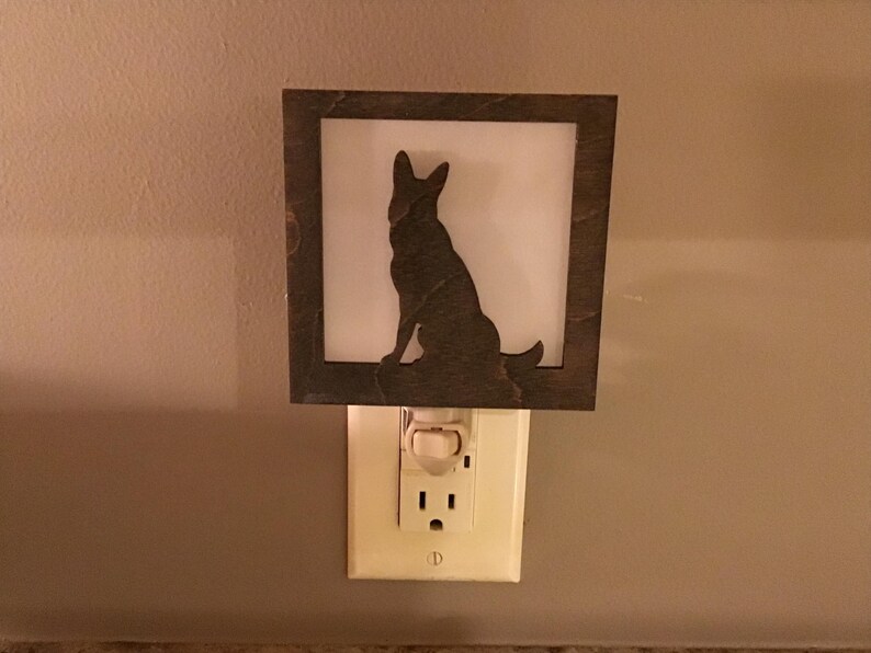 May include: A decorative night light featuring a silhouette of a dog within a square, dark-brown frame. The dog is sitting, facing right, against a white background. The night light plugs into a standard electrical outlet.