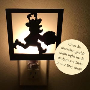 May include: A decorative night light featuring a black silhouette of a leprechaun running with a pot of gold. The night light is plugged into a wall outlet. Text on the image reads: Over 50 interchangeable night light shade designs available in our Etsy shop!