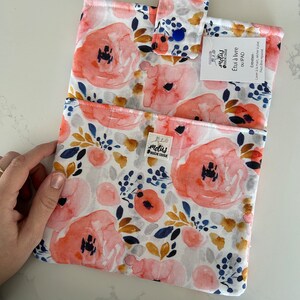 May include: iPad case with a floral watercolour design. The case features large coral-coloured flowers, navy blue berries, and grey accents on a white background. It has a tab closure with a blue button. The label reads "motus BOLDE COULE".